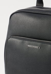 Sac en cuir noir texturé avec une fermeture éclair argentée et une plaque de logo en métal portant l'inscription "Armani Exchange". Surface lisse et forme structurée.