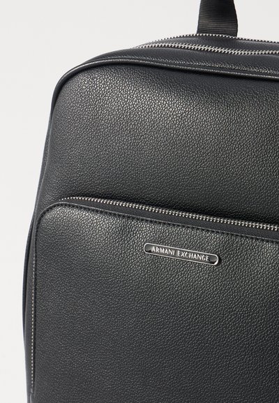 Sac en cuir noir texturé avec une fermeture éclair argentée et une plaque de logo en métal portant l'inscription "Armani Exchange". Surface lisse et forme structurée.