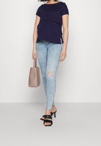 Blusa de maternidad marina con diseño en capas, jeans skinny rasgados azul claro, sandalias de tacón negras y un bolso tote beige. Look sencillo y contemporáneo.