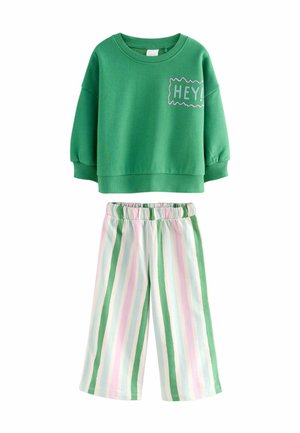 Grüner Langarmpullover mit dem Text "HEY!" kombiniert mit weißen, grünen, pinken und blauen vertikal gestreiften Gummizugbuxen.