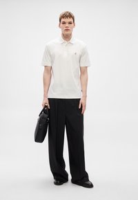 Λευκό πόλο μπλουζάκι με κοντά μανίκια, με μικρό λογότυπο στο στήθος, σε συνδυασμό με άνετα μαύρα παντελόνια και κρατώντας μια μαύρη τσάντα.