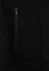 Tela acolchada negra con un bolsillo con cremallera prominente. Presenta una textura suave y patrones rectangulares uniformes en toda la superficie.