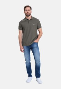Camisa polo de color verde oscuro con mangas cortas y tres botones, combinada con jeans de color azul claro y zapatillas blancas. Logo sutil en el pecho.