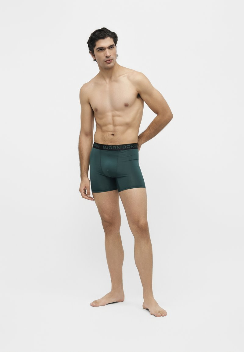 Homme torse nu debout pieds nus portant un boxer vert foncé avec une ceinture noire portant l'inscription Björn Borg sur fond blanc.
