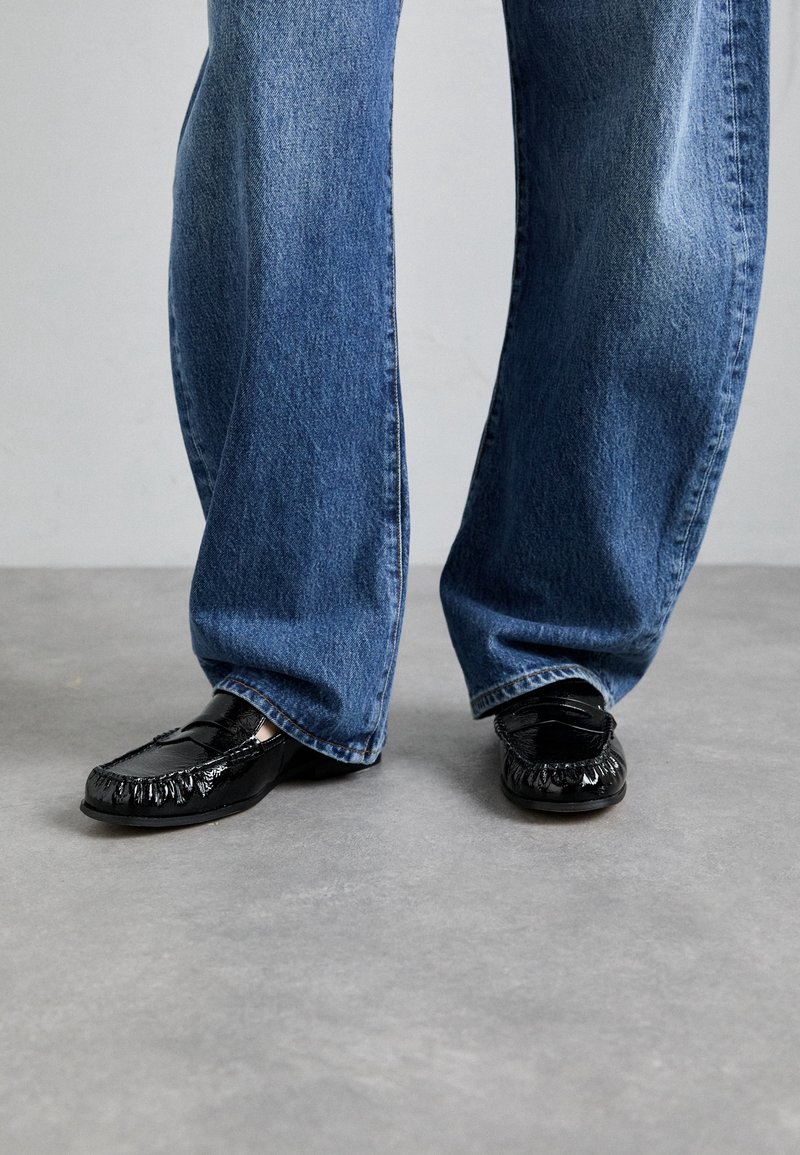 Jambes en jean bleu ample portant des mocassins noirs en cuir brillant, debout sur un sol gris avec un fond uni.