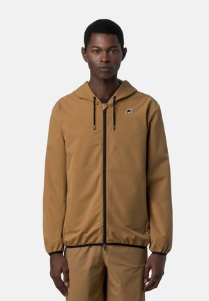 Uomo con giacca con cappuccio color beige con cerniera nera e pantaloni beige abbinati, in piedi davanti a uno sfondo grigio chiaro uniforme.