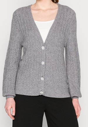 Femme portant un cardigan en maille grise texturée avec des boutons effet perle sur un haut blanc et un pantalon noir.