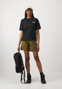 Musthave musta T-särk, millel on peegeldav plaastr, oliivrohelised cargo-lühikesed püksid ja must seljakott. Modell kannab kreemikat mütsi ja musti saapaid.