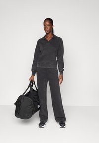 Felpa nera in pile con colletto, abbinata a pantaloni coordinati. Il modello tiene in mano una borsa da viaggio nera e indossa sneakers nere.