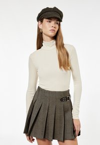 Helle beige Rollkragenoberteil mit langen Ärmeln, kombiniert mit einem plissierten braunen Hahnentrittröck mit einem Gürtelakzent. Accessoires mit einer strukturierten Mütze.