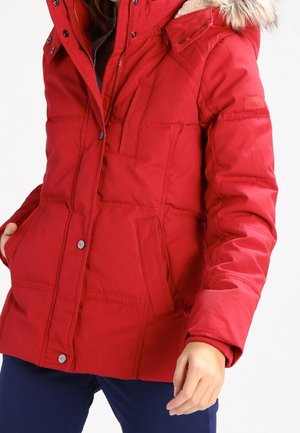 Kobieta ubrana w czerwoną zimową kurtkę z ociepleniem i kapturem obszytym futerkiem oraz granatowe spodnie, stojąca z jedną ręką w kieszeni.