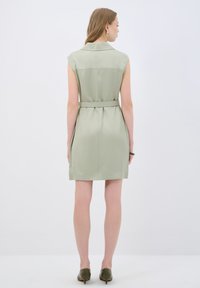 Robe sans manches, longueur genou, en tissu vert clair avec un col, une taille ceinturée et une drapé doux. Portée avec des chaussures vert olive.