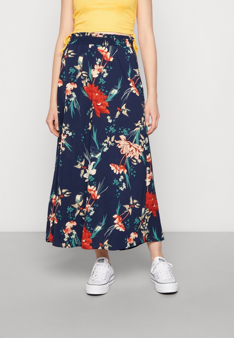 Femme portant une jupe midi fleurie bleu marine avec des fleurs rouges et beiges, un top jaune et des baskets blanches, debout sur un sol gris.
