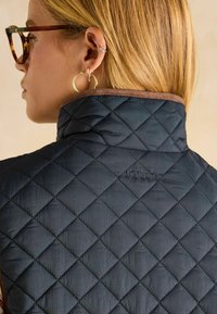 Pārsēta tumši zilā veste ar brūnu apdari, ar augstu apkakli un izšūtu "Joules" logo aizmugurē. Viegls audums ar gludu tekstūru.