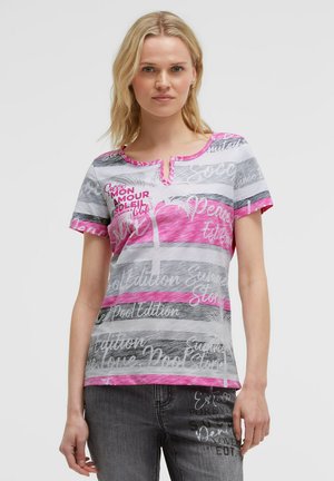 Blonde woman wearing a short-sleeve white and pink striped top with script text and dark gray jeans with white text designs.