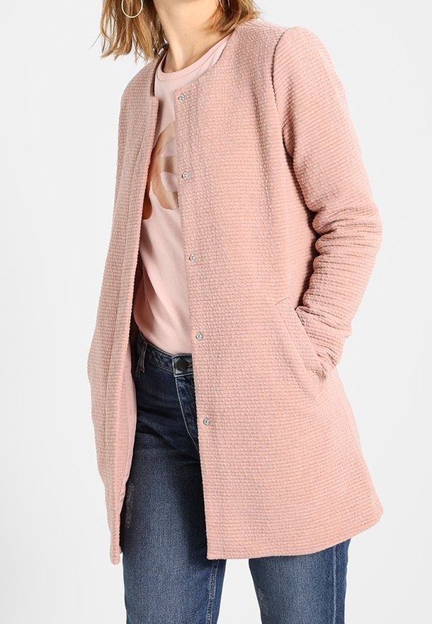 Rosa texturerad kappa med knäppning, långa ärmar och sidofickor, bärs över en ljusrosa skjorta och blå jeans.