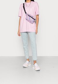 T-shirt oversize rosa con simbolo femminile, jeans dritti blu chiaro e sneakers nei colori pastello. La cintura a tracolla presenta un design con sfumatura viola.