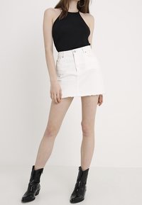 Kvinna i en ärmlös svart halternecktop, vit denimminikjol och svarta ankelboots, stående mot en helt vit bakgrund.