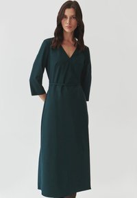 Robe enveloppante vert foncé avec décolleté en V, manches 3/4, lien à la taille et texture lisse, tombant sous le genou avec une coupe droite.
