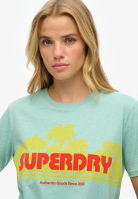 Mintgrøn bomulds t-shirt med et rødt "SUPERDRY" logo og gule palmetræsgrafikker, med rund hals og korte ærmer.