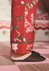 Țesătură roșie cu model floral care cade elegant, alături de pantofi slip-on negri cu detalii decorate și partea superioară transparentă.