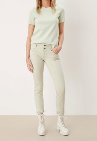 Ljusgrön t-shirt med kort ärm, i kombination med beiga åtsittande jeans och vita snörkängor. Noterbara detaljer inkluderar synlig sömnad och knappar.