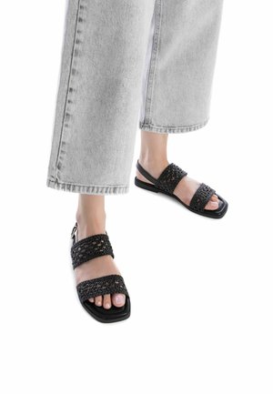 Sandalias negras trenzadas con dos correas anchas y punta cuadrada. Diseñadas con una suela plana y correa para el tobillo ajustable. Llevadas con unos vaqueros claros recortados.
