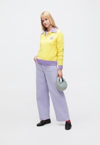 Pull-over en tricot jaune avec un col violet, associé à un pantalon large en velours côtelé violet clair. Le mannequin porte un sac à main en tissu gris.