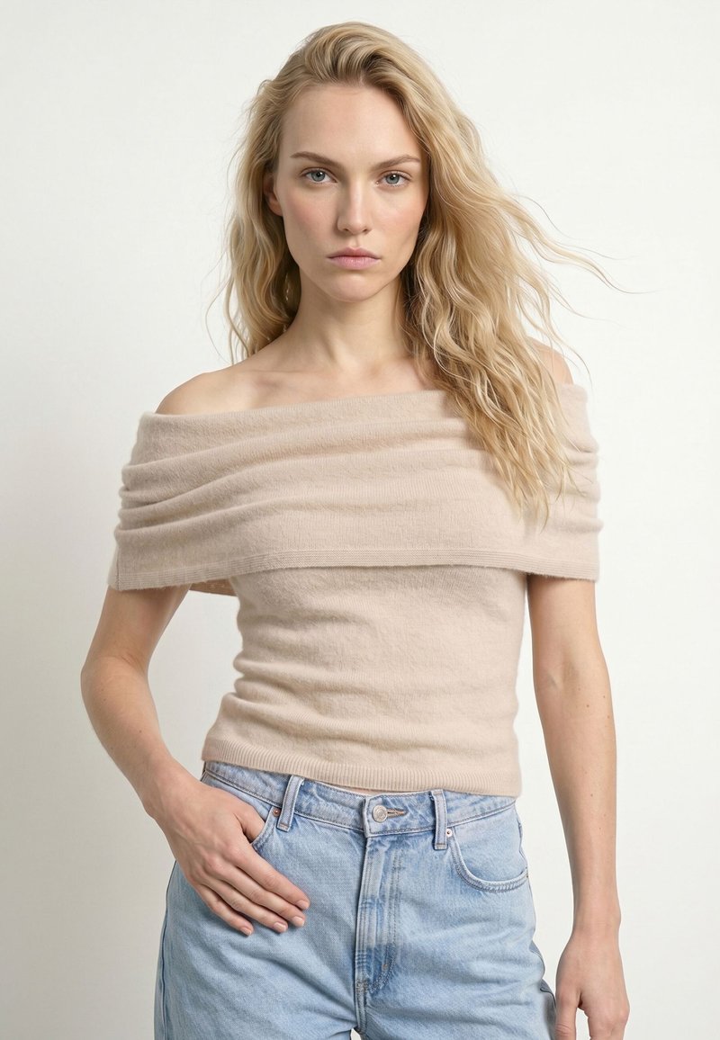 Jeune femme aux longs cheveux blonds porte un haut beige décolleté aux épaules et un jean bleu clair, debout devant un fond blanc uni.