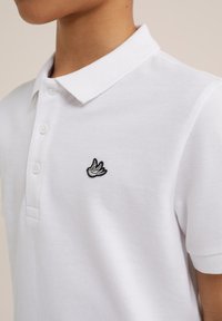 Baltas polo marškinėlis, pagamintas iš tekstūrinio audinio, su dviem mygtukais. Krūtinėje yra mažas juodas ir baltas siuvinėtas paukščio logotipas.