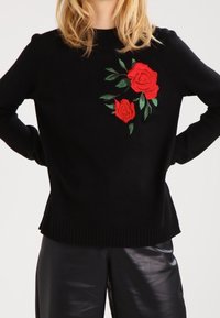 Czarny sweter dzianinowy z haftem czerwonej róży na lewym ramieniu, z prążkowanymi mankietami i dolnym wykończeniem, połączony z błyszczącymi czarnymi skórzanymi spodniami.