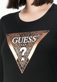 T-shirt nera con un grande triangolo in rame metallico e stampa leopardata che presenta "GUESS U.S.A" e un logo centrale a forma di punto interrogativo.