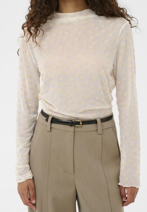 Mujer que lleva un top blanco transparente de manga larga con puntos texturizados, pantalones beige de talle alto y un cinturón negro con hebilla dorada.