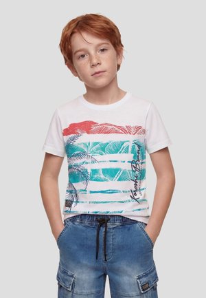 Niño pelirrojo con pecas vestido con una camiseta blanca con estampado tropical y pantalones cortos cargo azules, de pie con las manos en los bolsillos contra un fondo gris.