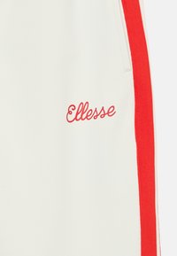 Krémové nohavice s červenými bočnými pruhmi a vyšitým červeným logom "Ellesse" na ľavom stehne. Hladká textúra s športovým dizajnom.