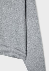 Manche de pull en laine gris clair et détail du bord-côtes sur un fond clair uni.