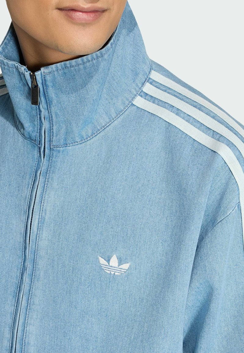 Man draagt een lichtblauwe denim jas met rits, voorzien van wit Adidas-logo en drie witte strepen op de schouder.