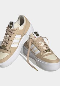 Beige ja valkoinen alustasneakerit, joissa on paksut valkoiset pohjat, beige narut, Adidas-logot kielellä ja kolme valkoista raitaa sivuilla.