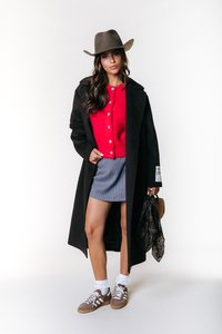 Lang svart frakk over en rød cardigan, stripete skjørt og brune sneakers med hvite sokker. Modellen har på seg en bredbrimmet brun hatt og holder en veske.