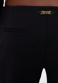 Calças pretas ajustadas com uma textura suave, um pequeno bolso traseiro e um detalhe em metal dourado com o logo da Versace Jeans Couture.