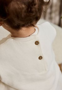 Kinderpullover in Creme mit geripptem Rundhalsausschnitt und dreiknöpfigem Verschluss an der Rückseite. Weiche Textur und schlichtes Design.