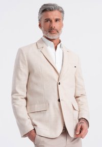 Blazer de lino beige con solapa muestrera, dos bolsillos frontales y botones marrones. Llevado sobre una camisa blanca con cuello. Textura suave.