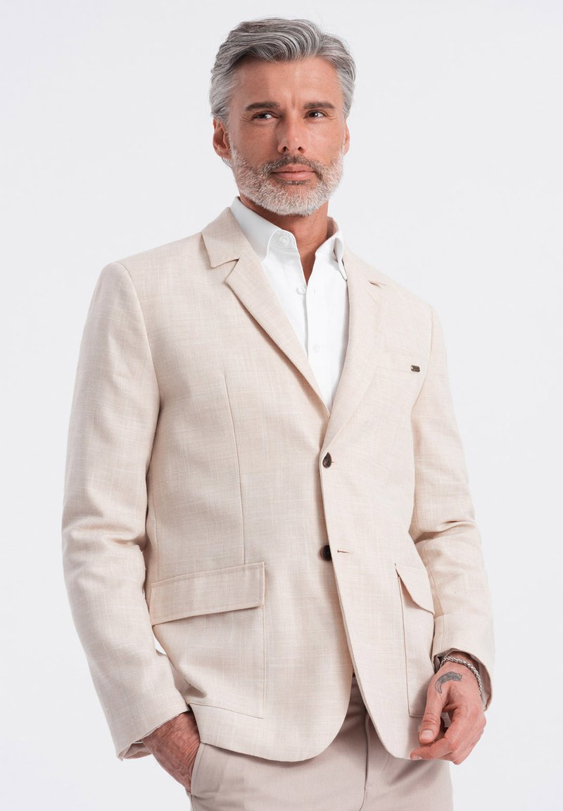 Blazer de lino beige con solapa muestrera, dos bolsillos frontales y botones marrones. Llevado sobre una camisa blanca con cuello. Textura suave.