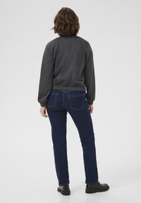 Femme aux cheveux bruns mi-longs portant une veste gris foncé, un jean bleu et des chaussures noires, debout de dos sur un fond blanc.