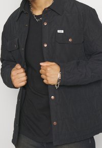 Black quilted jacket with snap buttons, two front pockets, and a fabric label. Worn over a black shirt, featuring a textured surface.