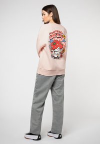 Helles pinkes Sweatshirt mit einem bunten Grafikdesign eines lächelnden Charakters, der eine Erdbeere hält, kombiniert mit grauen Hosen und schwarzen Sneakers.