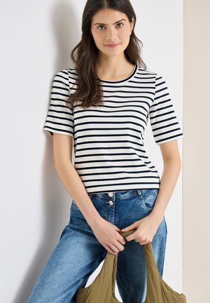 Ung kvinde iført en sort-hvid striped kortærmet skjorte og blå jeans, der holder en grøn mulepose med begge hænder.