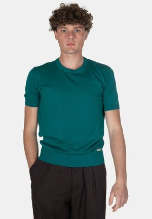 Giovane uomo con capelli ricci che indossa un maglione a maniche corte color verde acqua e pantaloni scuri, in piedi con una mano in tasca su sfondo bianco.