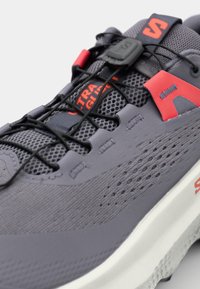 Szare buty sportowe z siateczkowymi i syntetycznymi materiałami, teksturowanymi wzorami, czerwonymi akcentami, elastycznymi sznurówkami i białą podeszwą.
