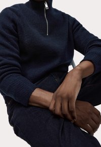 Granatowy, strukturalny sweter z dzianiny z wysokim kołnierzem i zamkiem błyskawicznym do połowy, w połączeniu z ciemnymi dżinsami, eksponującymi ściągacze przy mankietach.